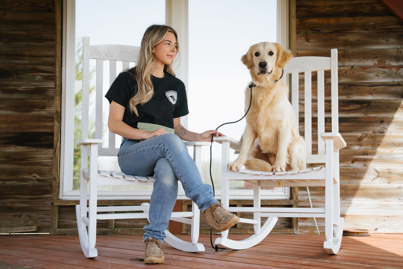 A trainer and a golden retriever sit in white rocking chairs on a rustic wood porch