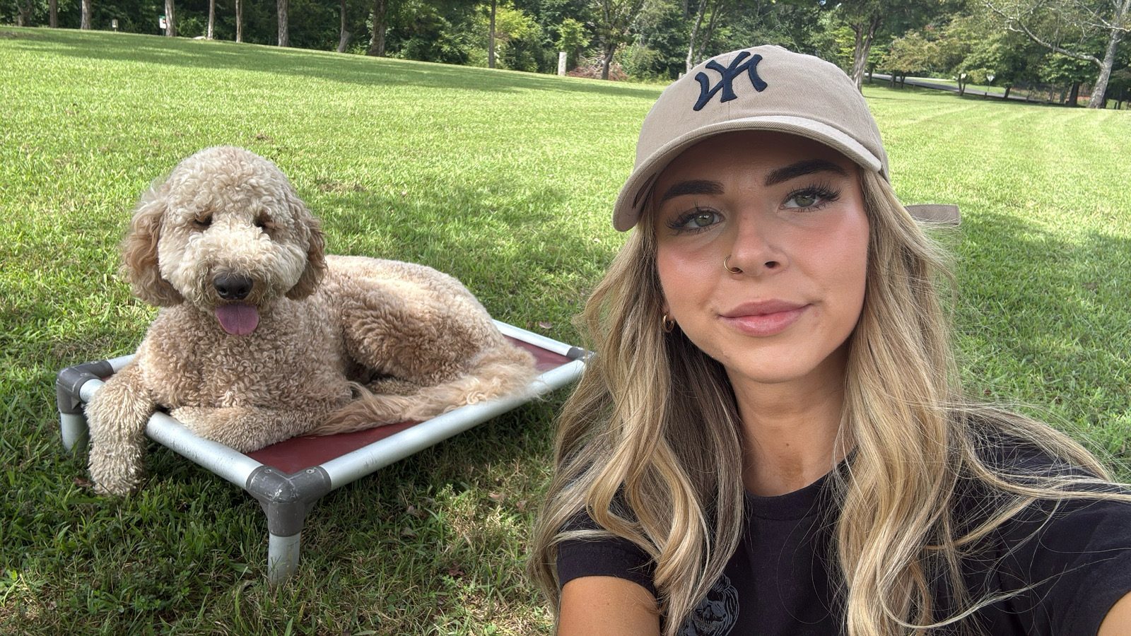 A trainer takes a selfie beside a curly goldendoodle lying on a raised dog bed on the lawn