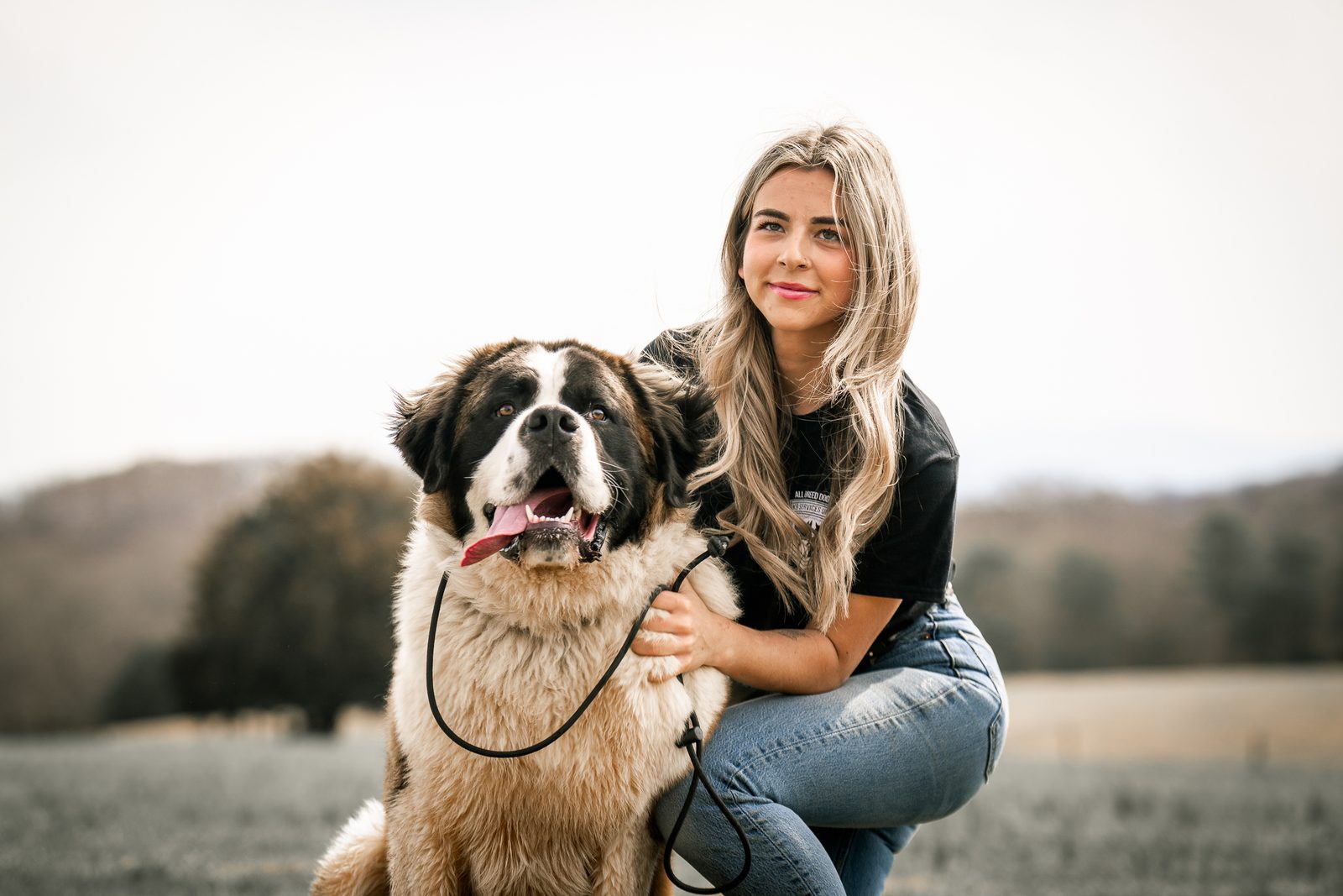 A trainer kneels with her arm around a smiling Saint Bernard in a hilltop field